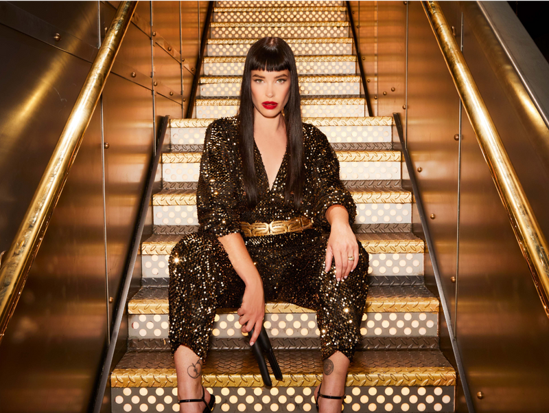 model sitting on gold and silver stairs wearing a sparkling sequins jumpsuit with a gold belt holding a CLOUD NINE Original Cordless Iron 