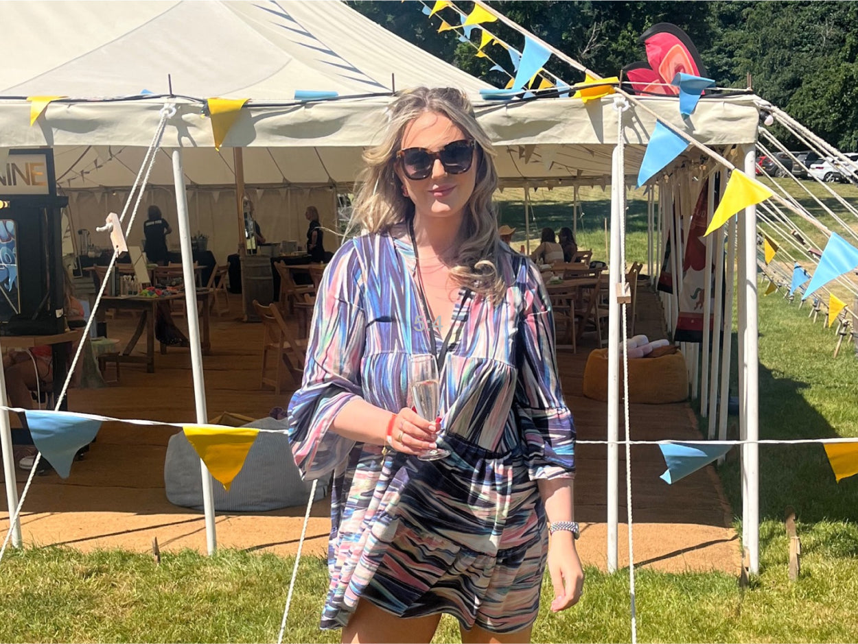 picture of a female CLOUD NINE employee in a artsy designed dress with black sunglasses on with a drink in her hand with a marquee behind her with blue and yellow flags