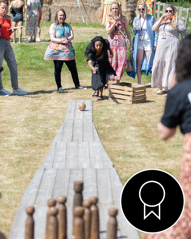 woman mid throw playing bowling with wooden pins and ball down a wooden lane while people watch 