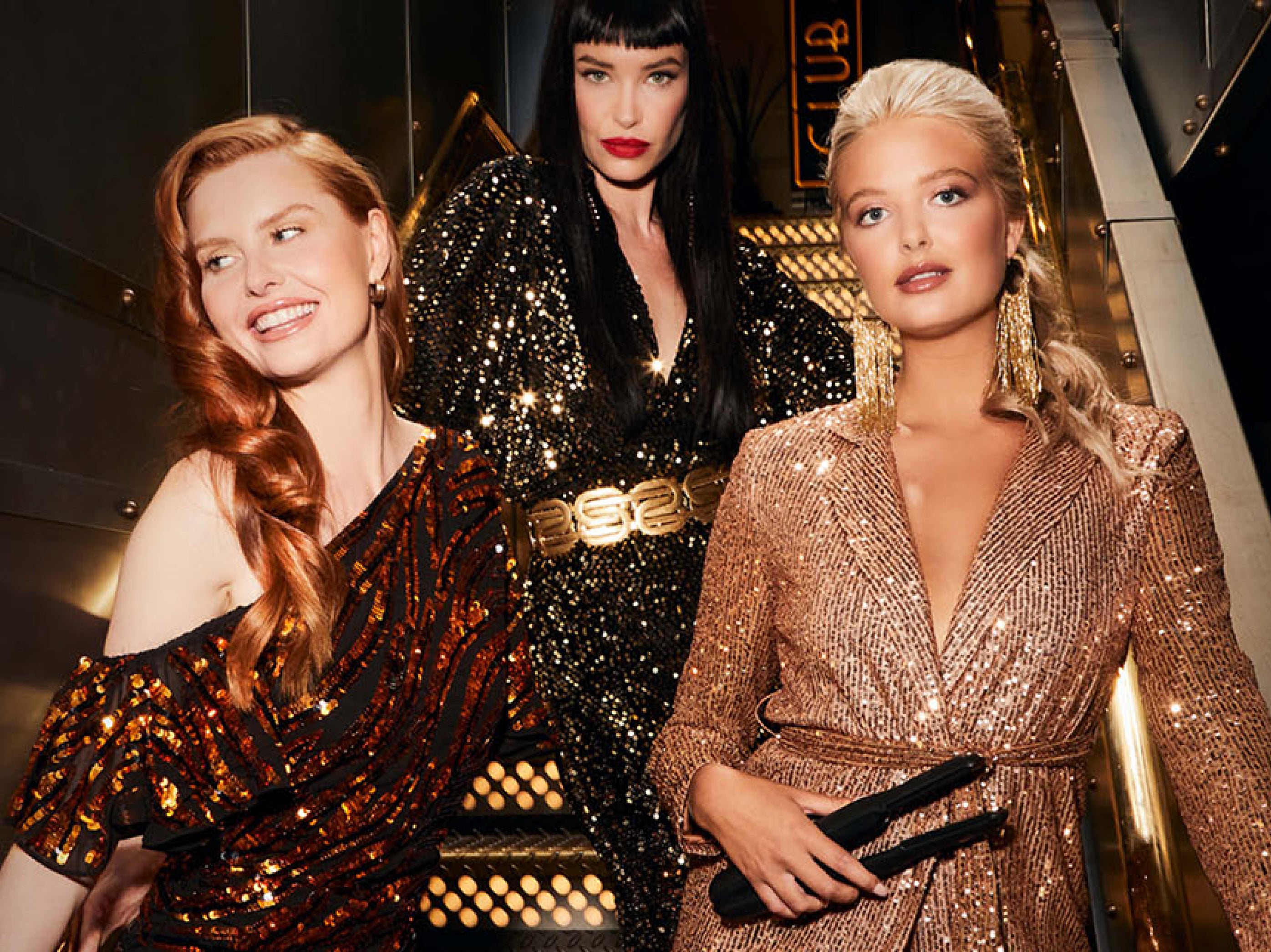 three models, one ginger, one jet black hair, one blonde in sequined outfits posing on a staircase 